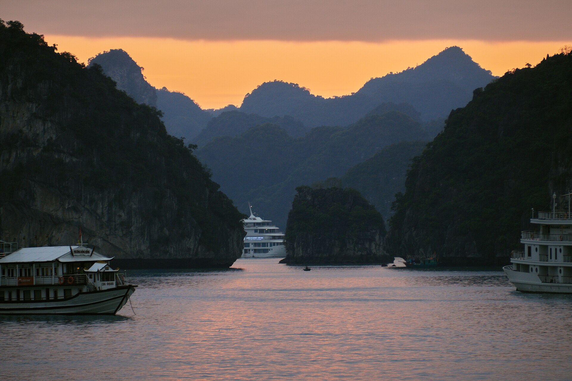 Overnight cruise boats anchored among limestone islands in Ha Long Bay under an orange evening sky