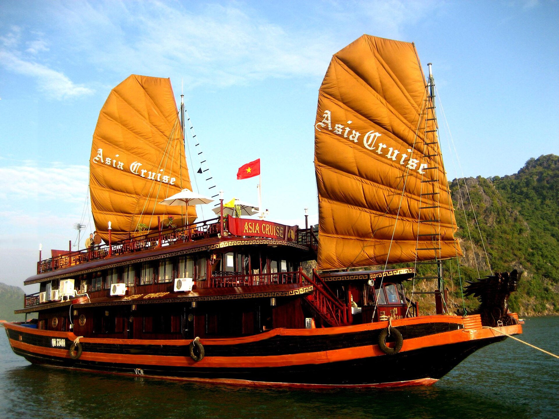 Best Ha Long Bay Cruises: How to Choose (Not a Top 10) 2 A traditional-style wooden junk with two ochre sails and a dragon-head prow moored near a wooded limestone island