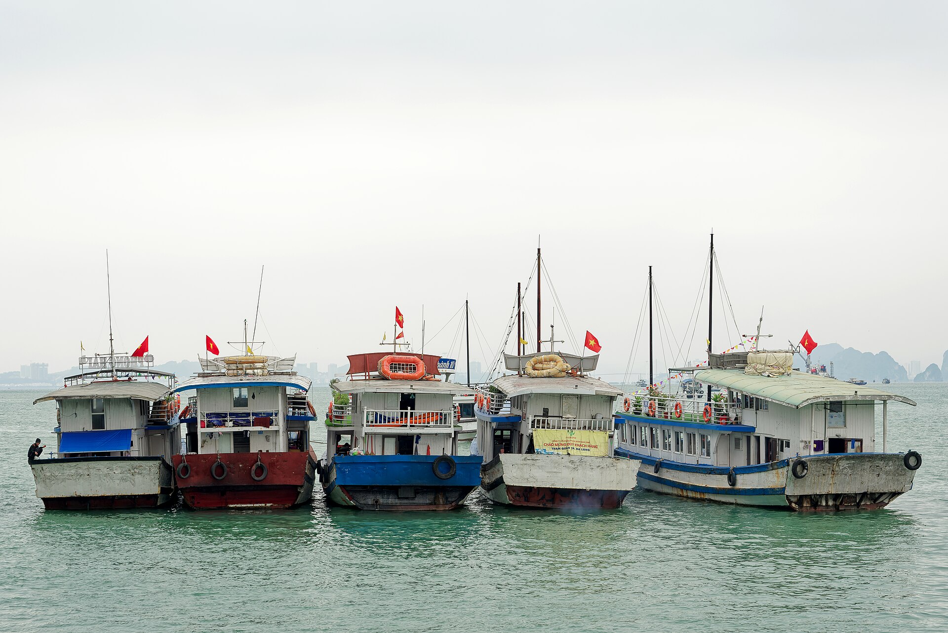 Ha Long Bay 2 Day 1 Night: What the Itinerary Actually Looks Like 2 Five tourist boats with Vietnamese flags moored side by side off Ti Top Island on a grey day